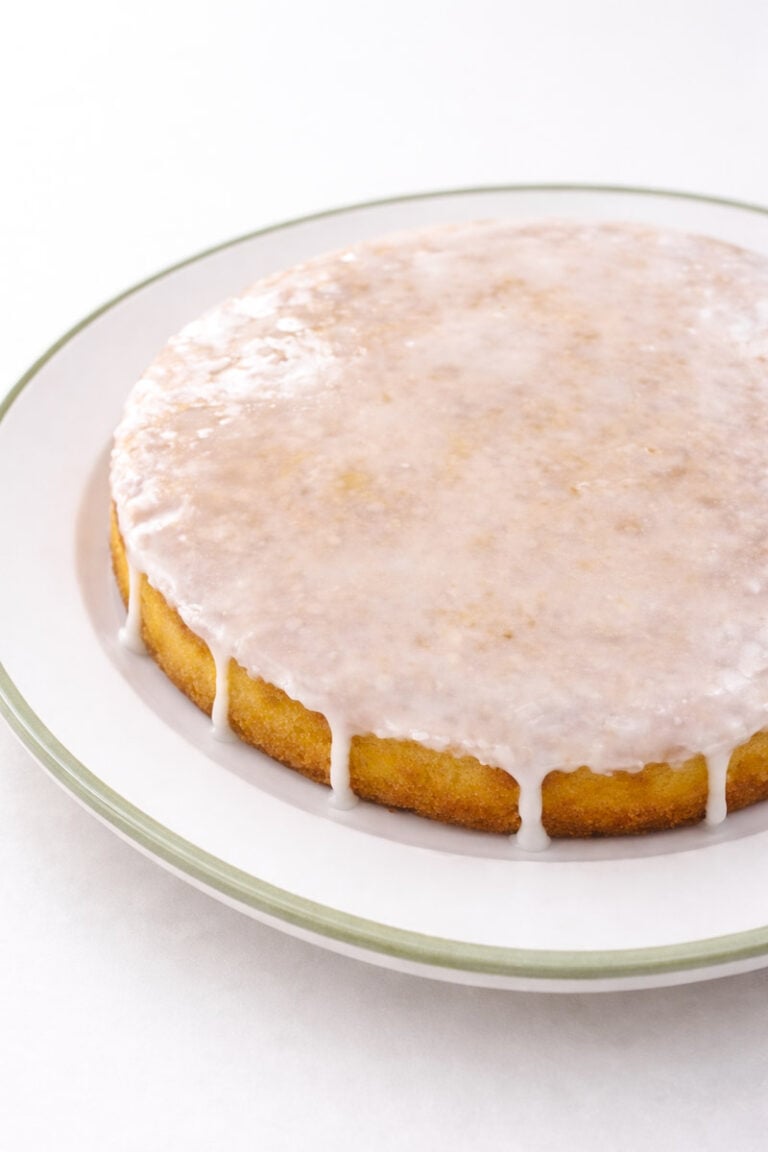 A round, golden lemon cake sits on a white plate with a green rim, topped with a smooth layer of white glaze that drips slightly over the edges.