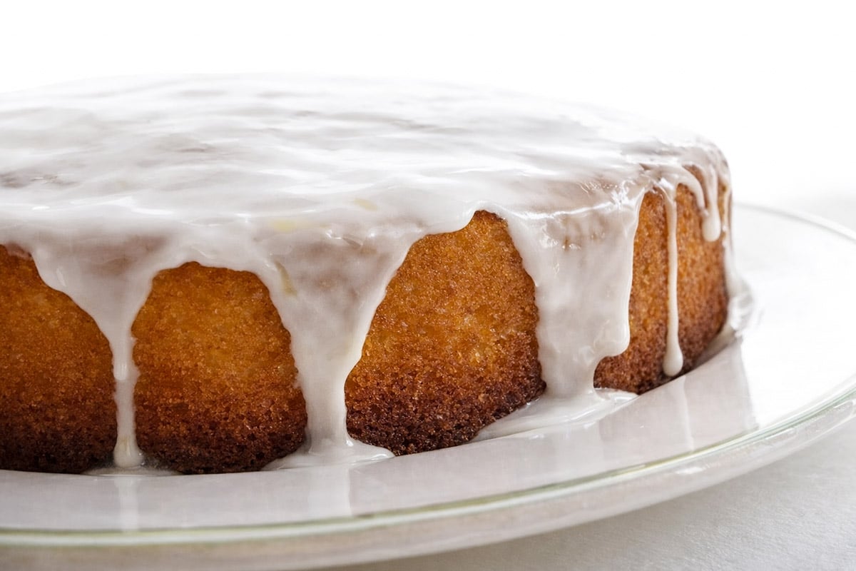A round, golden-brown lemon cake sits on a glass plate, topped with a thick layer of white icing that drips down the sides. The cake rests on a white surface with a softly lit background.