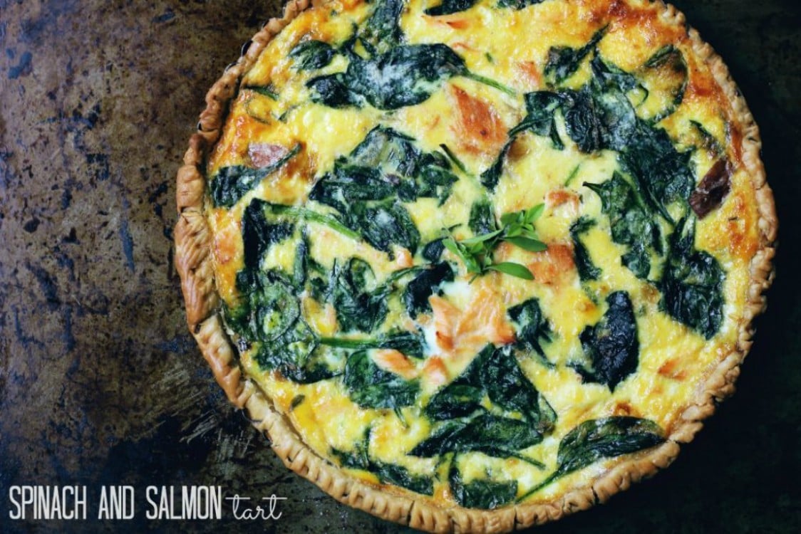 A baked tart with a golden crust, filled with spinach, salmon chunks, and melted cheese, displayed on a rustic dark surface. Text in the corner reads SPINACH AND SALMON QUICHE.
