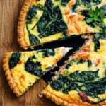 A salmon quiche with spinach and a golden crust sits on a wooden surface. One slice is slightly pulled out, revealing the creamy filling with tender pieces of salmon and vibrant green spinach leaves.