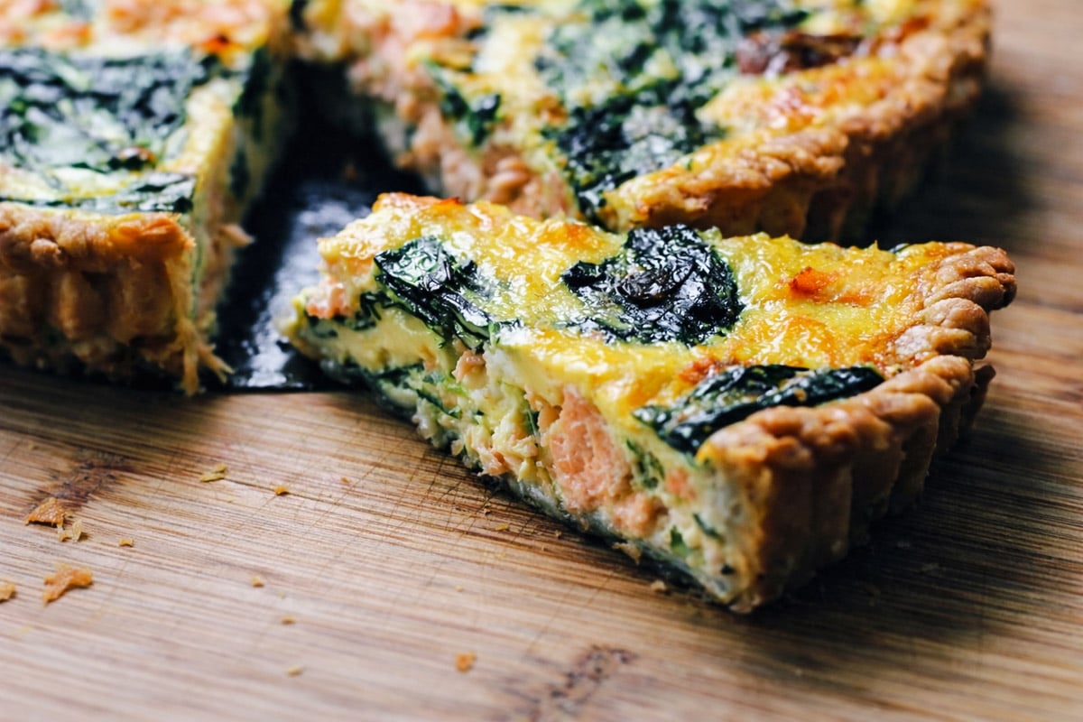 A close-up of a sliced salmon quiche on a wooden cutting board, featuring a golden crust and creamy filling with visible spinach leaves and chunks of salmon.