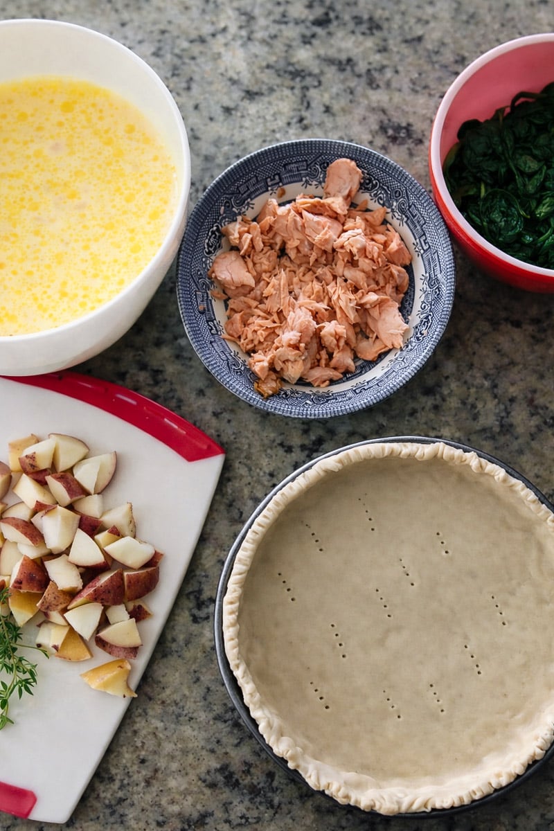 Overhead view of ingredients for a salmon quiche: a bowl of beaten eggs, cooked salmon, cooked spinach, chopped red potatoes on a cutting board, and an unbaked pie crust in a pan.