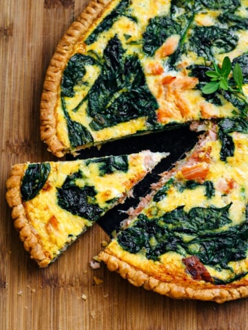 A salmon quiche with spinach and a golden crust sits on a wooden surface. One slice is slightly pulled out, revealing the creamy filling with tender pieces of salmon and vibrant green spinach leaves.