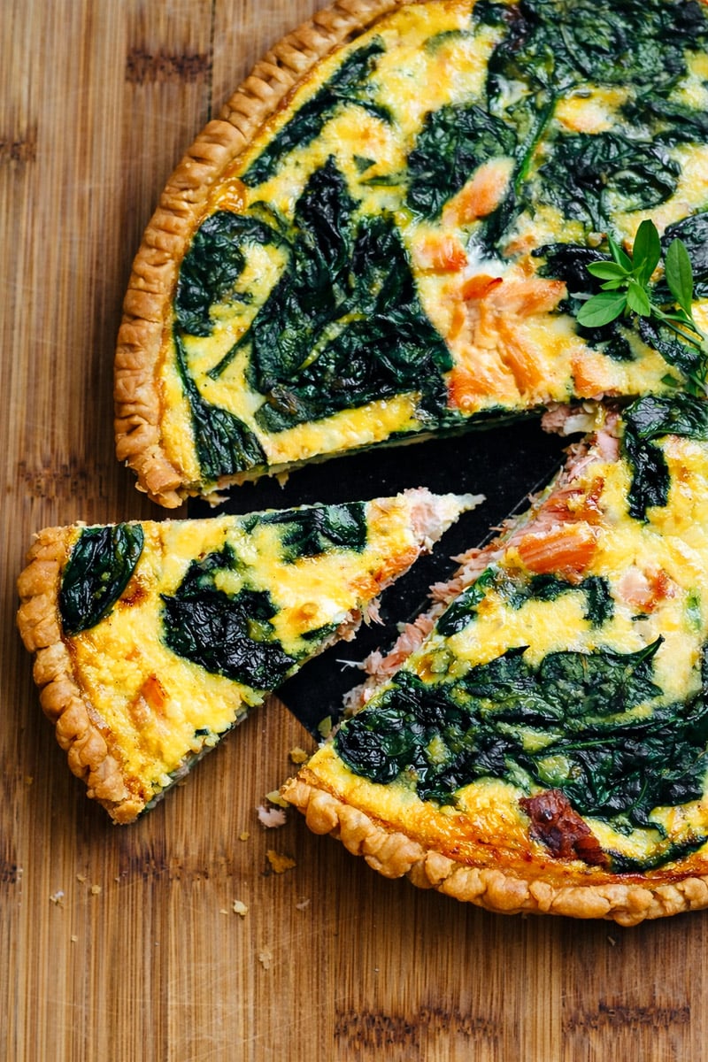 A salmon quiche with spinach and a golden crust sits on a wooden surface. One slice is slightly pulled out, revealing the creamy filling with tender pieces of salmon and vibrant green spinach leaves.