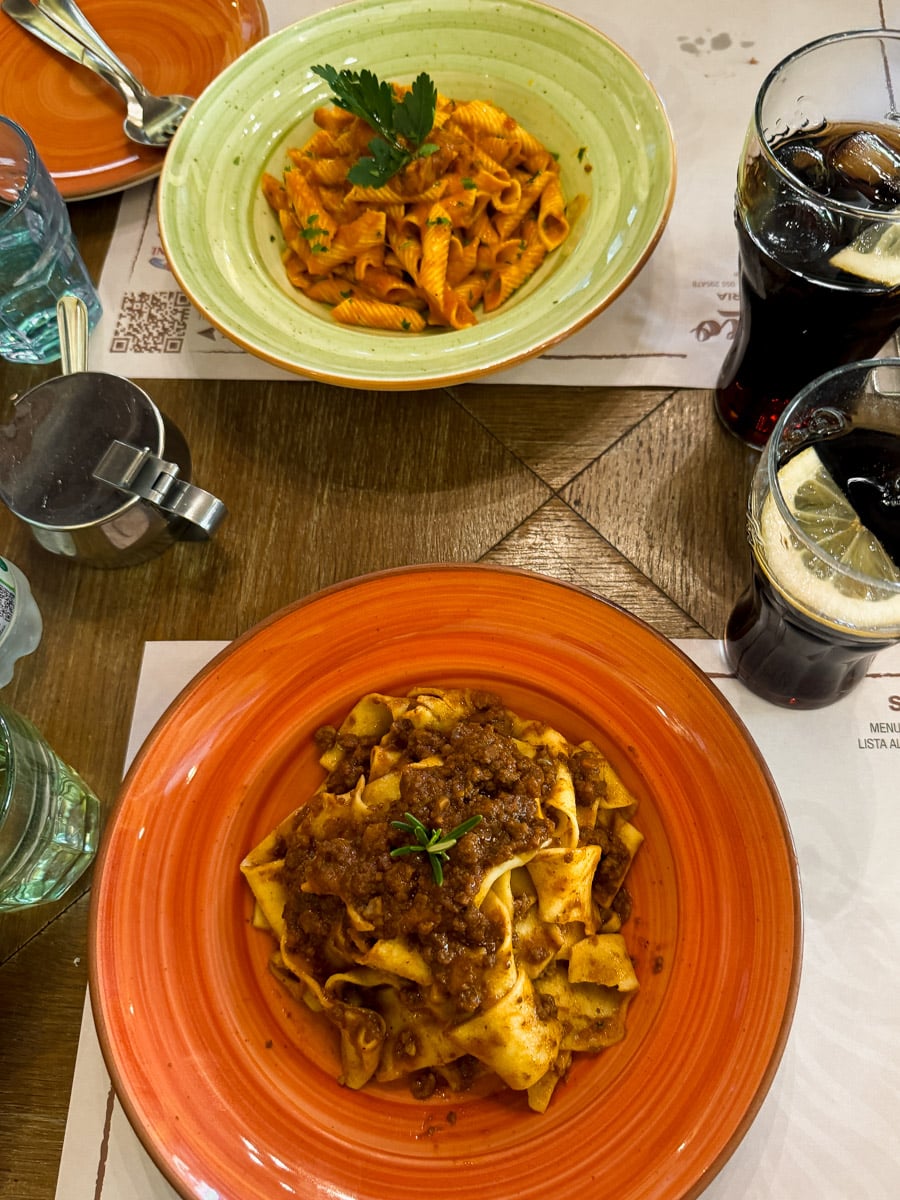 Two plates of pasta on a wooden table in Siena, Italy: one with ribbon pasta in meat sauce, the other with short pasta in red tomato sauce. Two glasses of cola with ice and water complete this classic Siena dining experience.