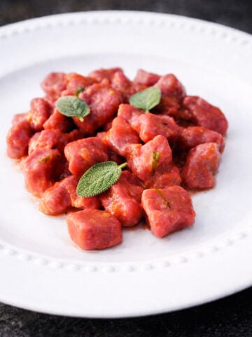 A white plate with a serving of vibrant beet and potato gnocchi garnished with fresh sage leaves, set against a dark background.