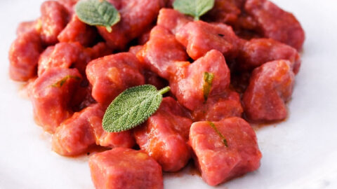 A white plate with a serving of vibrant beet and potato gnocchi garnished with fresh sage leaves, set against a dark background.