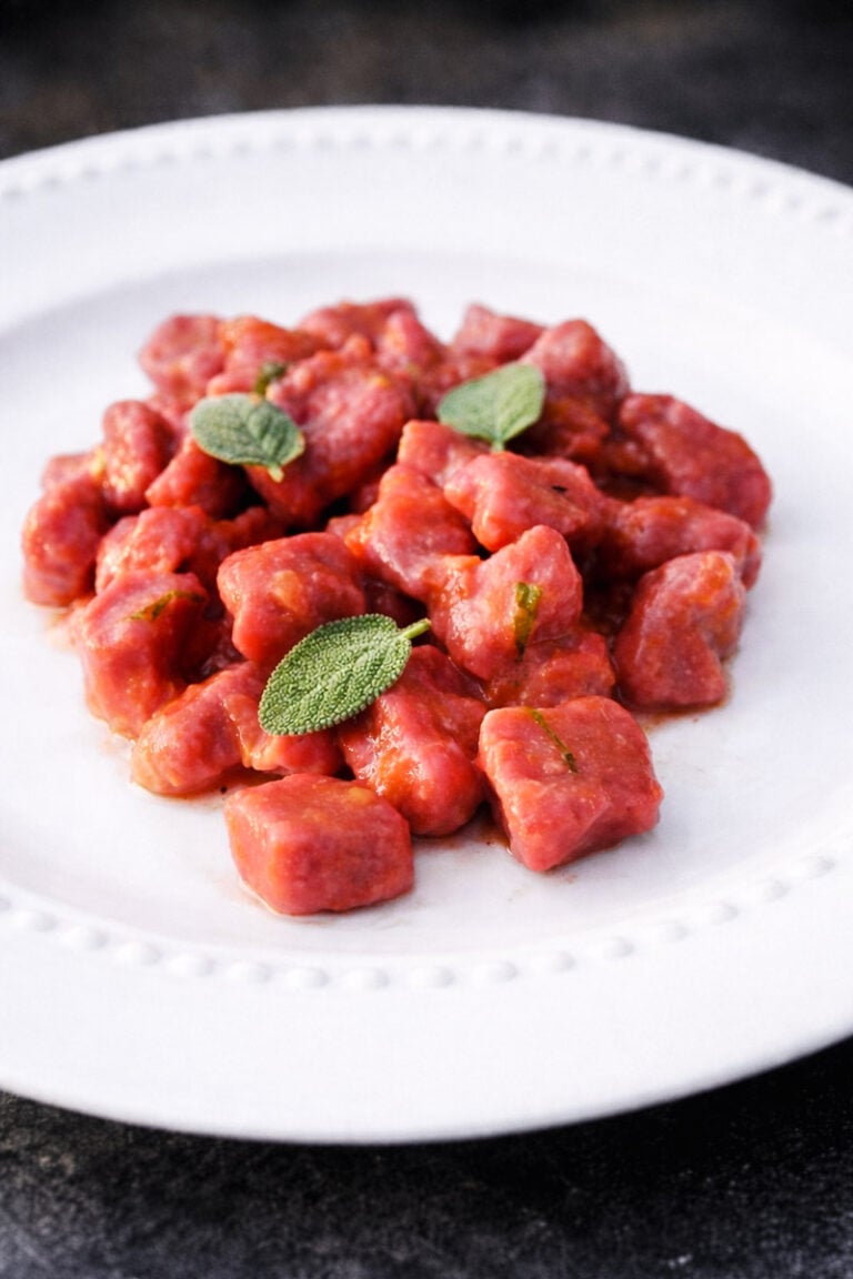 A white plate with a serving of vibrant beet and potato gnocchi garnished with fresh sage leaves, set against a dark background.