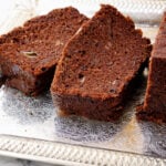 Three slices of moist Chocolate Date Bread are arranged on an ornate silver tray atop a white marble surface. The bread boasts a rich, dark brown color with visible crumbs and delicious chunks nestled inside.