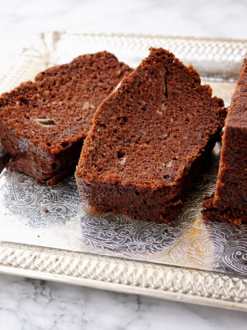 Three slices of moist Chocolate Date Bread are arranged on an ornate silver tray atop a white marble surface. The bread boasts a rich, dark brown color with visible crumbs and delicious chunks nestled inside.