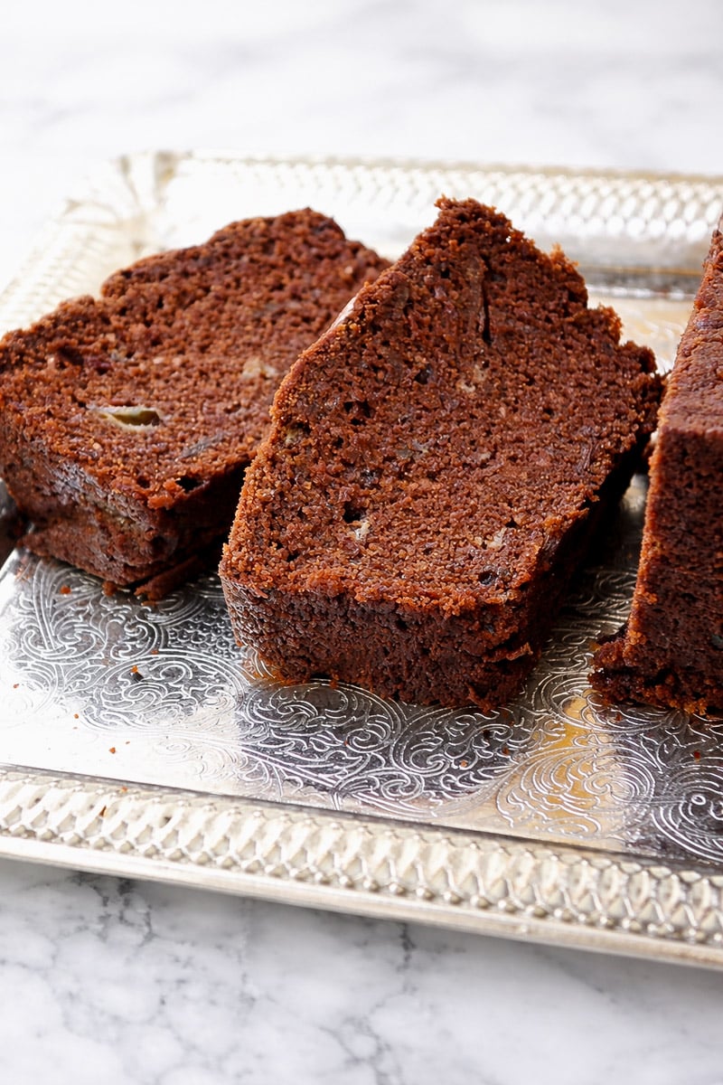 Three slices of moist Chocolate Date Bread are arranged on an ornate silver tray atop a white marble surface. The bread boasts a rich, dark brown color with visible crumbs and delicious chunks nestled inside.