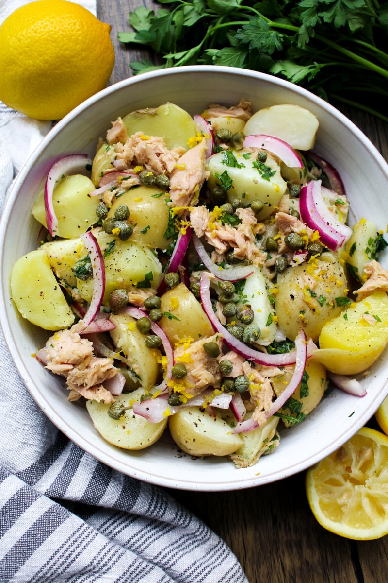 close up image of tuna and potato salad in a white bowl with parsley and lemon in background.