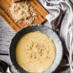 A bowl of creamy, pale yellow soup topped with grated cheese sits on a rustic wooden table. Beside it, a wooden board holds a block of cheese and a pile of freshly grated cheese, with a cloth and spoon nearby.