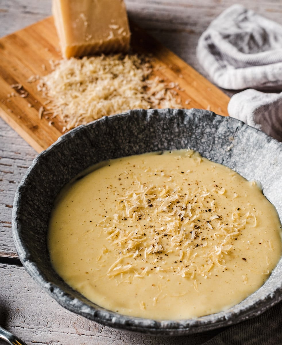 A gray bowl filled with creamy polenta topped with grated cheese and black pepper sits on a rustic wooden table; a wooden board with more grated cheese and a chunk of parmesan is in the background.