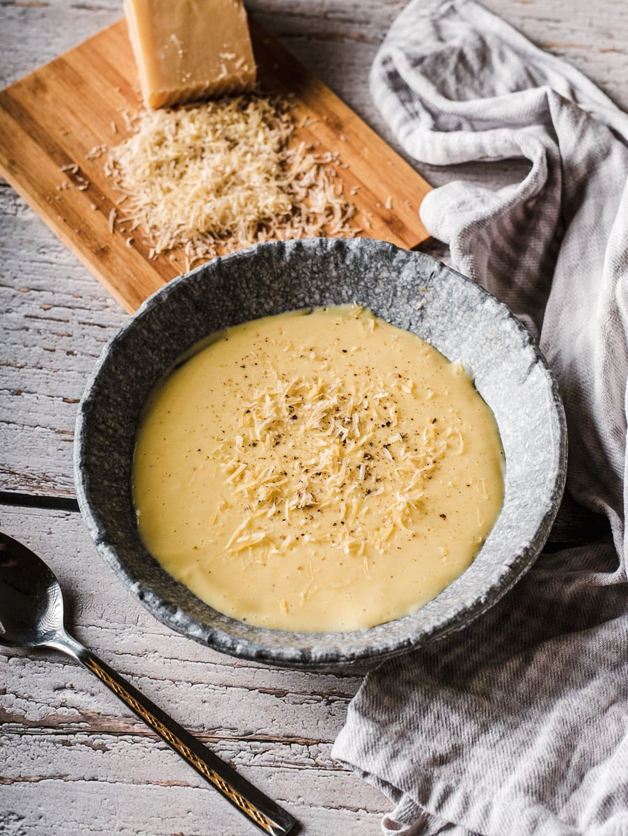 A bowl of creamy soup topped with grated cheese sits on a rustic wooden table, next to a spoon, a gray striped cloth, and a wooden board with a wedge of cheese and grated cheese on it.