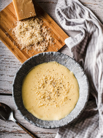 A bowl of creamy, pale yellow soup topped with grated cheese sits on a rustic wooden table. Beside it, a wooden board holds a block of cheese and a pile of freshly grated cheese, with a cloth and spoon nearby.