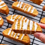 A hand holding a piece of easy pumpkin spice biscotti with white icing drizzled on top; more iced biscotti rest on a cooling rack in the background.