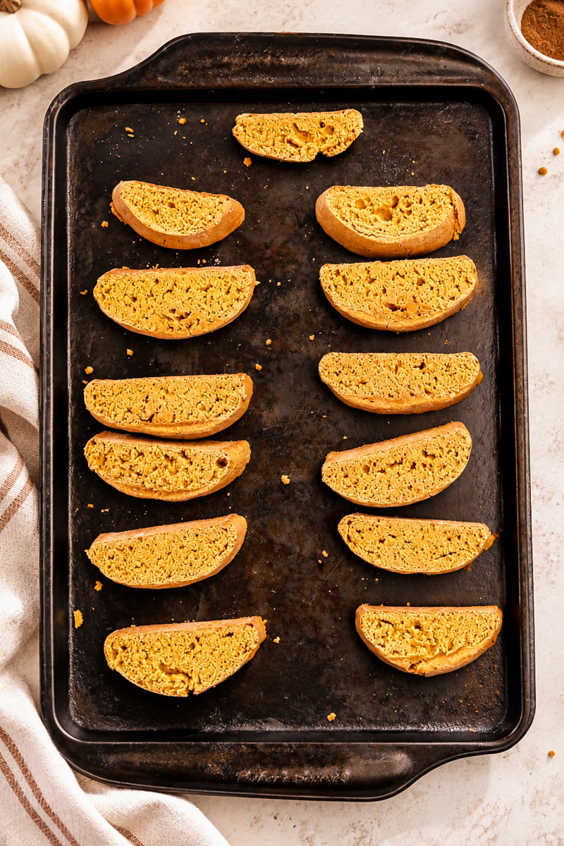Twelve easy pumpkin spice biscotti slices arranged in neat rows on a dark baking tray, with a striped towel on the side and a light countertop background.