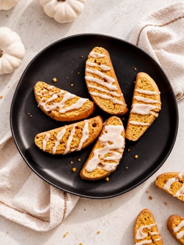 Six easy pumpkin spice biscotti with white icing drizzle rest on a black plate, surrounded by tan cloth napkins and small white decorative pumpkins on a light surface.