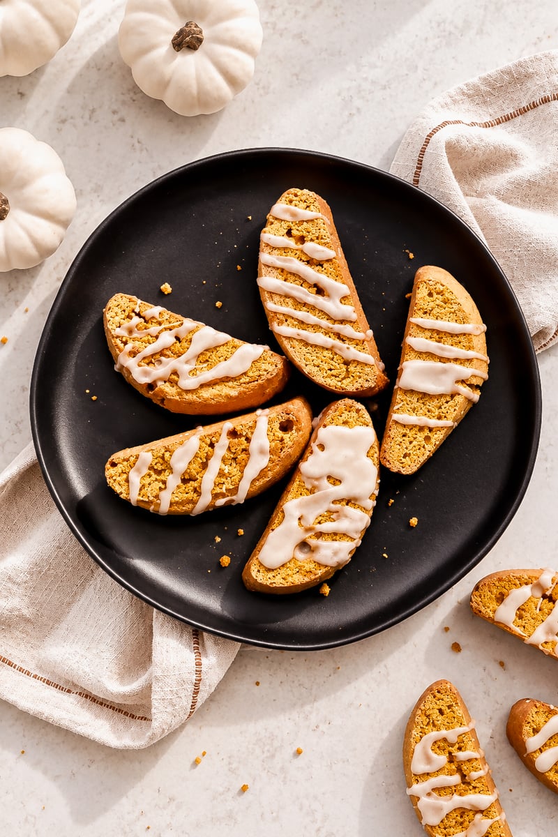 Six easy pumpkin spice biscotti with white icing drizzle rest on a black plate, surrounded by tan cloth napkins and small white decorative pumpkins on a light surface.