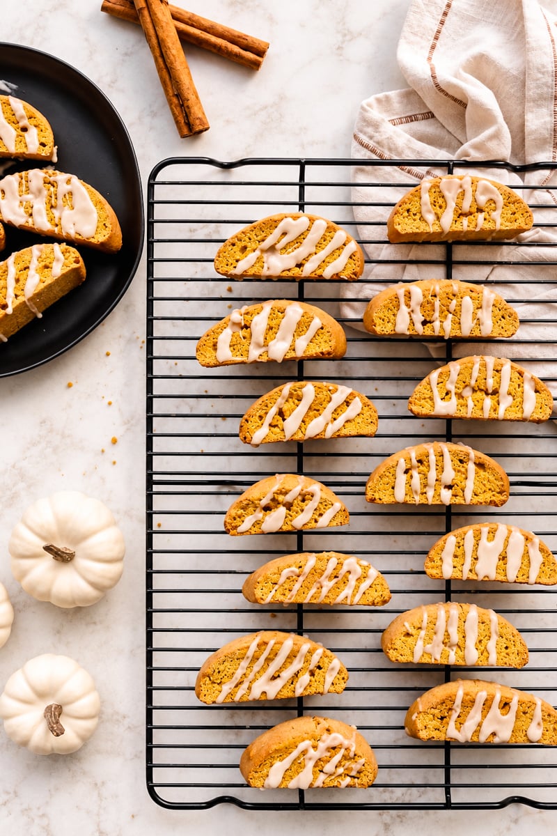 Easy pumpkin spice biscotti with white icing are arranged on a wire cooling rack. A plate with more biscotti, white pumpkins, cinnamon sticks, and a striped kitchen towel sit nearby on a marble surface.
