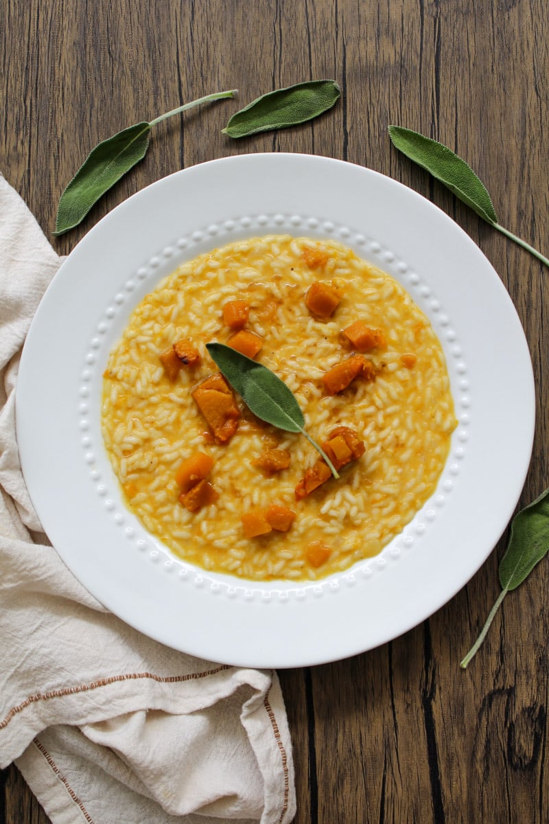 A white plate of creamy risotto with chunks of butternut squash, garnished with a sage leaf, sits on a wooden table with a beige napkin and scattered sage leaves.