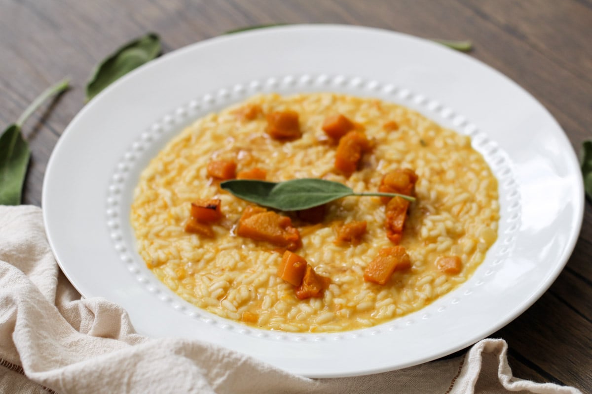 A white plate holds creamy risotto topped with chunks of orange squash and garnished with a fresh sage leaf, set on a wooden table with a beige napkin nearby.