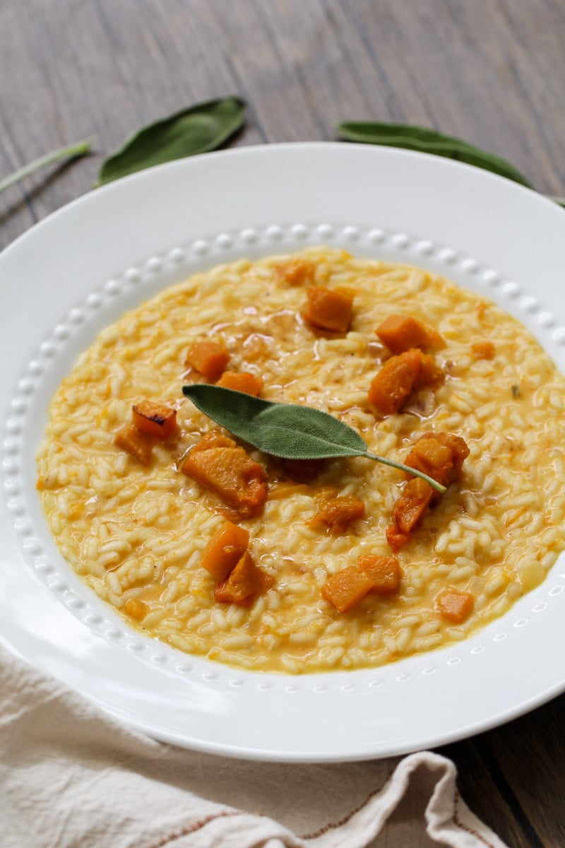 A white plate of creamy risotto topped with golden butternut squash cubes and garnished with a fresh sage leaf, placed on a wooden surface with a napkin partially visible.