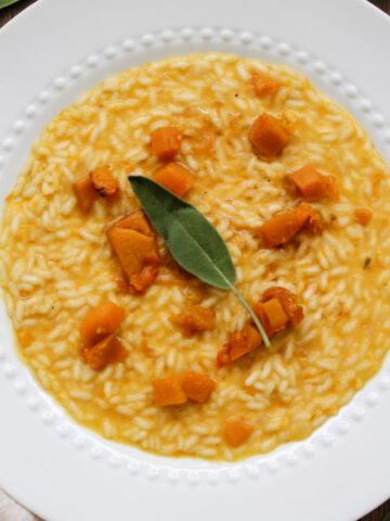 A white plate with creamy risotto topped with chunks of roasted butternut squash and garnished with a fresh sage leaf.
