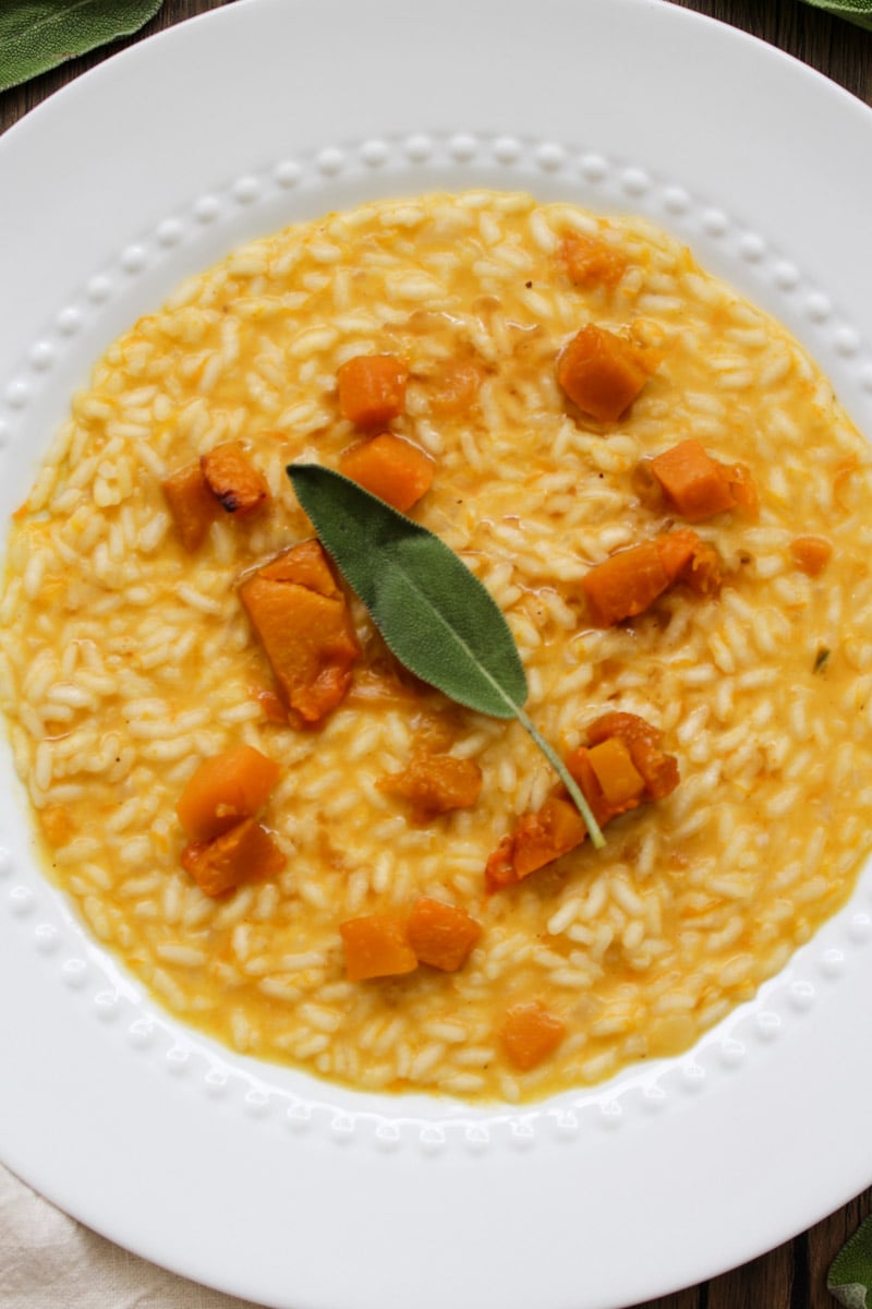 A white plate with creamy risotto topped with chunks of roasted butternut squash and garnished with a fresh sage leaf.
