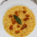 A white plate of creamy butternut squash risotto, garnished with roasted squash cubes and a fresh sage leaf, sits on a wooden table with a striped cloth and additional sage leaves beside it.