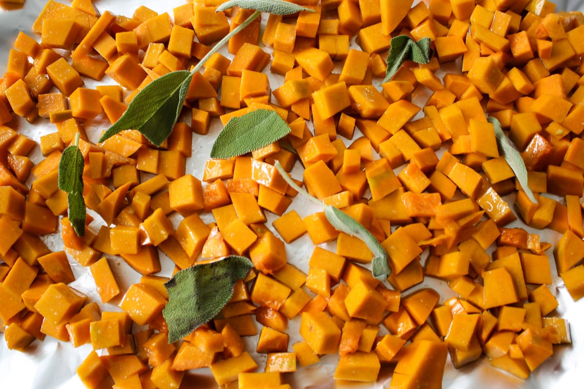 Diced butternut squash and whole sage leaves spread out on a white surface, ready for roasting.