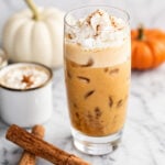 Iced pumpkin spice latte with whipped cream in a glass, topped with cinnamon. Two cinnamon sticks, a white pumpkin, an orange pumpkin, and a mug of creamy pumpkin spice latte are in the background on a marble surface.