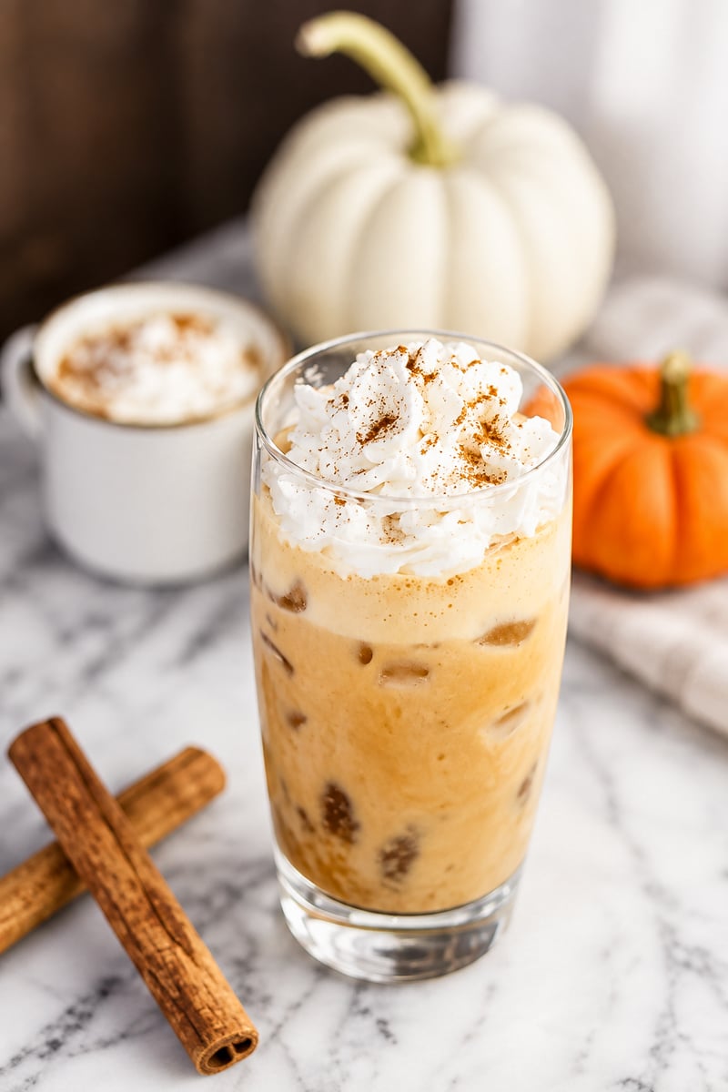 Iced pumpkin spice latte topped with whipped cream and a sprinkle of cinnamon, on a marble surface with two cinnamon sticks, a small white pumpkin, an orange pumpkin, and a cup of coffee in the background—a perfect autumn-inspired pumpkin spice latte scene.