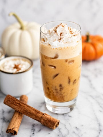 Iced pumpkin spice latte with whipped cream in a glass, topped with cinnamon. Two cinnamon sticks, a white pumpkin, an orange pumpkin, and a mug of creamy pumpkin spice latte are in the background on a marble surface.