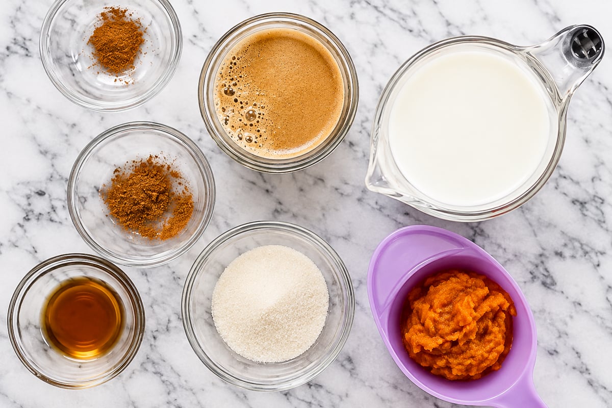 Ingredients on a marble counter evoke the cozy essence of a pumpkin spice latte: ground spices, brewed espresso, milk in a glass jug, vanilla extract, sugar, and pumpkin puree in a purple bowl.