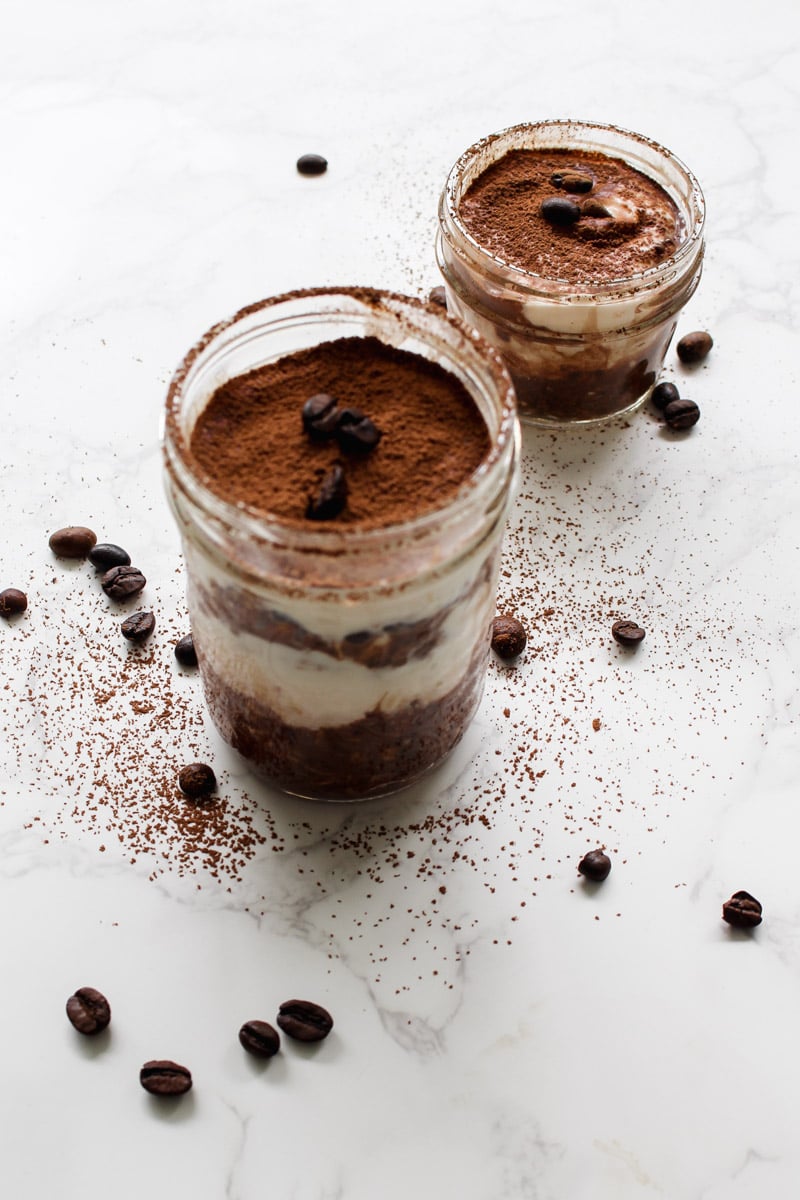 Two glass jars filled with layered tiramisu, topped with cocoa powder and coffee beans, sit on a white marble surface with coffee beans and cocoa powder scattered around.