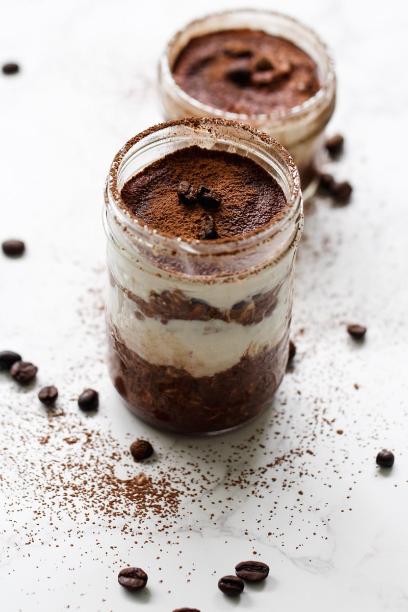 Two glass jars filled with layered tiramisu overnight oats, topped with cocoa powder and coffee beans, sit on a white surface sprinkled with cocoa and scattered coffee beans.