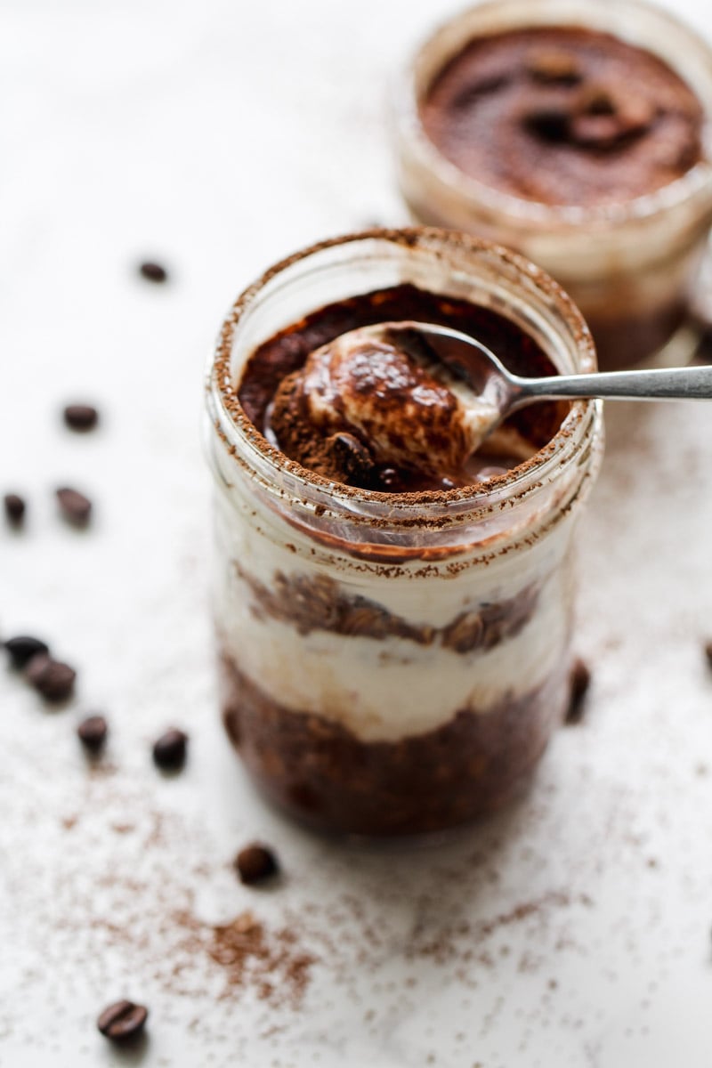 A close-up of a jar filled with layered tiramisu, topped with cocoa powder. A spoon is scooping out a portion. Coffee beans and cocoa powder are scattered on the white surface around the jar.