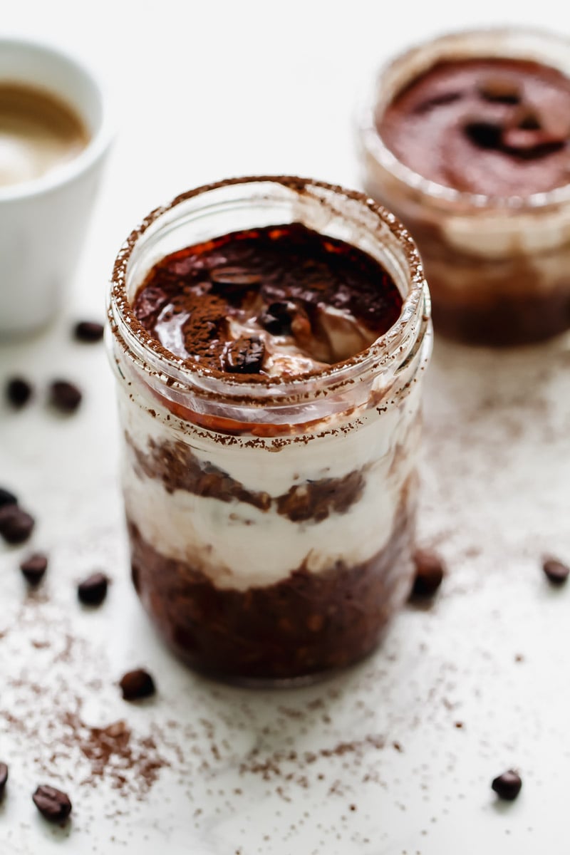 A close-up of a layered dessert in a glass jar with chocolate, cream, and cocoa powder, surrounded by coffee beans and a cup of coffee in the background. Another jar is slightly blurred behind it.