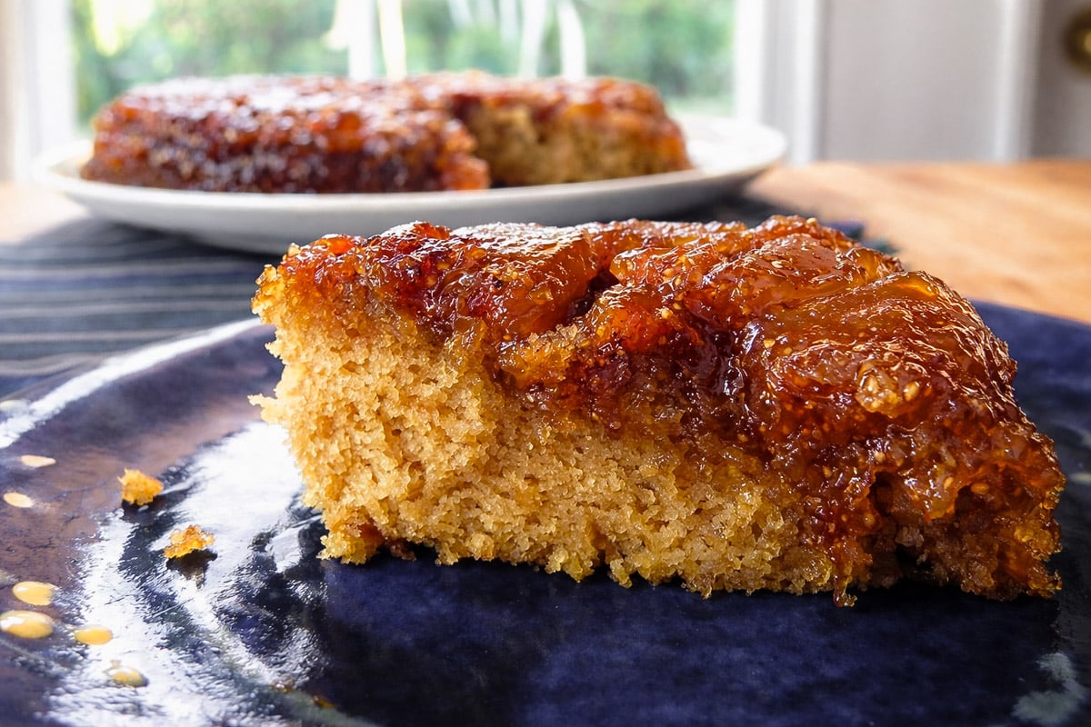 A close-up of a slice of vegan fig upside-down cake on a blue plate, with another larger piece on a white plate in the background. The cake appears moist and golden, topped with glossy, caramelized figs.