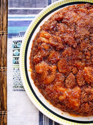 A close-up of a round, caramelized vegan fig upside-down cake with glistening golden-brown fruit topping, served on a white plate with a green and black rim, placed on a striped tablecloth.