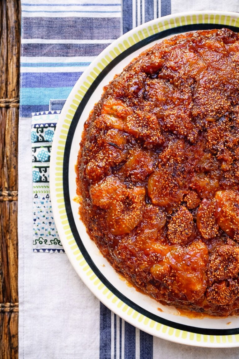 A close-up of a round, caramelized vegan fig upside-down cake with glistening golden-brown fruit topping, served on a white plate with a green and black rim, placed on a striped tablecloth.