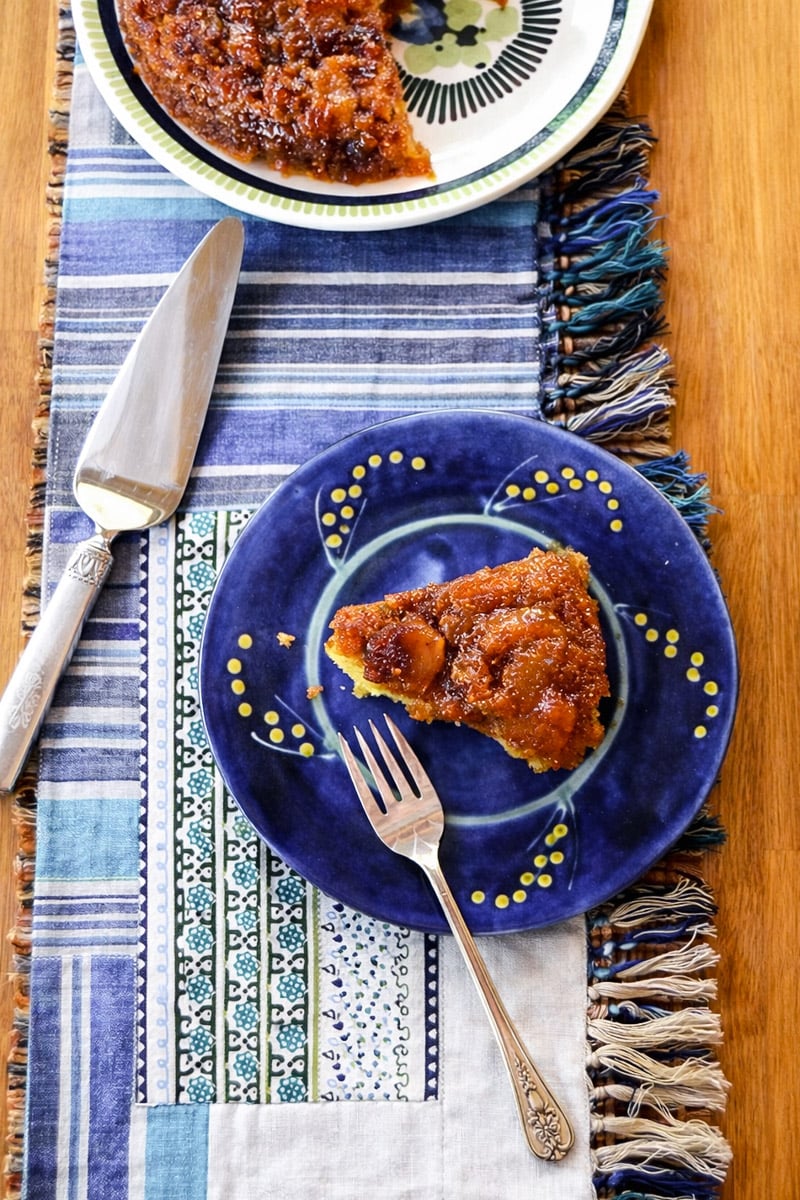 A slice of vegan fig upside-down cake sits on a blue plate with a fork, next to a pie server and another plate on a patterned, fringed table runner atop a wooden table.