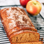 A loaf of spiced apple bread, reminiscent of an Apple Cider Donut Cake and dusted with sugar, sits on a cooling rack with two slices cut. Two apples are in the background on a marble surface.