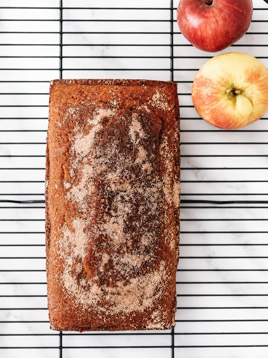 A rectangular loaf of apple bread with a cinnamon-sugar topping, reminiscent of an Apple Cider Donut Cake, sits on a black cooling rack beside two red-yellow apples on a white marble surface.