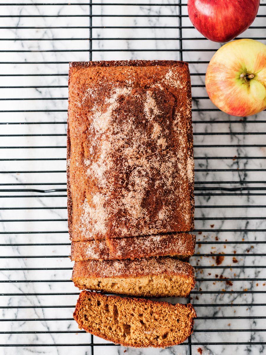 A loaf of apple bread with a cinnamon-sugar topping sits on a cooling rack, reminiscent of the cozy flavors found in an Apple Cider Donut Cake. Two slices are cut, and two apples are placed nearby on a marble surface.