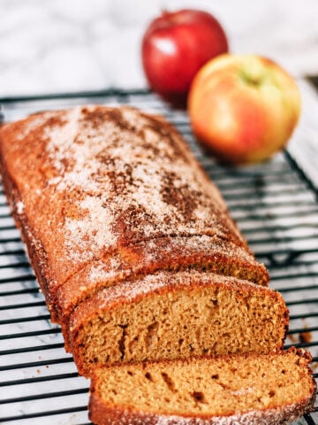 A loaf of spiced apple bread, reminiscent of an Apple Cider Donut Cake and dusted with sugar, sits on a cooling rack with two slices cut. Two apples are in the background on a marble surface.