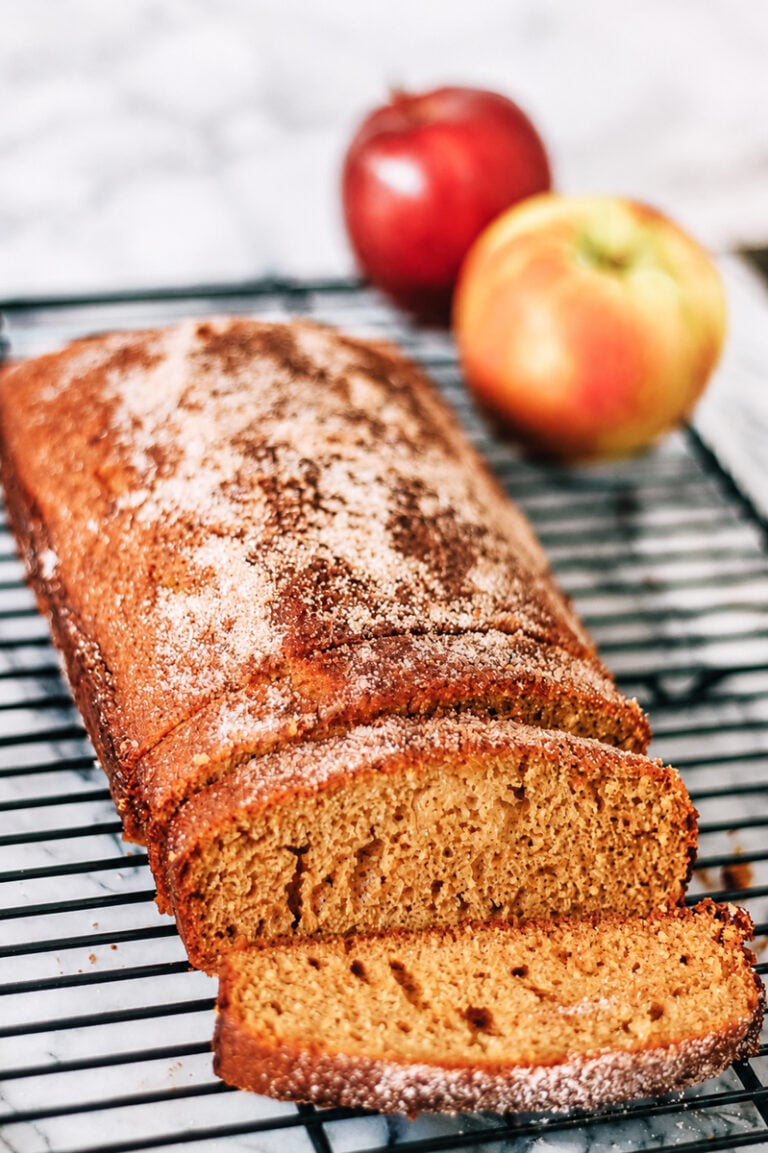 A loaf of spiced apple bread, reminiscent of an Apple Cider Donut Cake and dusted with sugar, sits on a cooling rack with two slices cut. Two apples are in the background on a marble surface.