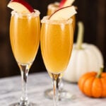 Three champagne flutes filled with a golden Apple Cider Mimosa cocktail, garnished with apple slices and sugar rims, sit on a marble surface with white and orange pumpkins in the background.